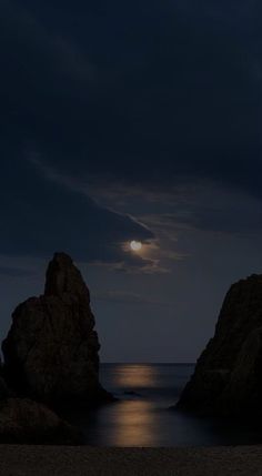 the moon is setting over the ocean with rocks in the foreground and dark blue sky