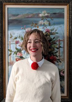 a woman wearing a white sweater with a red pom - pom in front of a painting