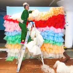 a woman on a ladder painting the side of a multi - colored wall with layers of tulle