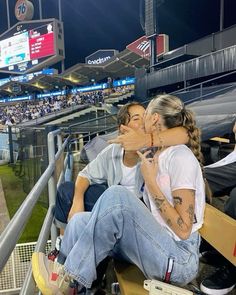 two people sitting in the bleachers at a baseball game, one kissing the other