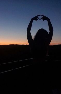 a woman holding up two hands in the shape of a heart at sunset or dawn