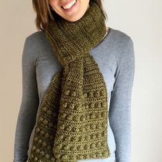 a smiling woman wearing a green crocheted scarf and grey shirt with her hands in her pockets