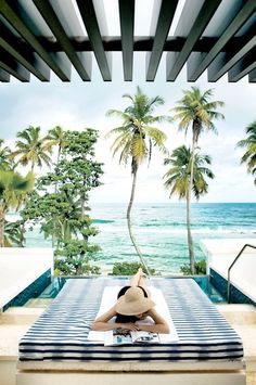 a woman laying on top of a bed in front of palm trees and the ocean