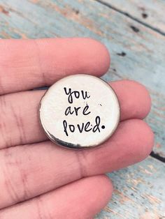 a hand holding a small button with the words you are loved written in black ink