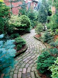 a brick path in the middle of a garden with trees and shrubs on either side