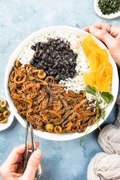 two hands holding spoons over a bowl filled with rice, beans and other foods