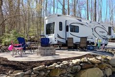 a camper parked in the woods next to a stone wall with chairs around it