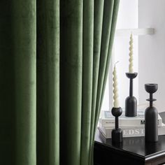 two candles are sitting on a table next to a window with green curtains and books