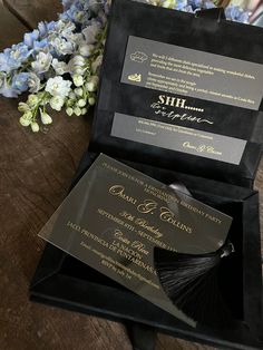 a black and gold wedding card in a box next to blue flowers on a wooden table