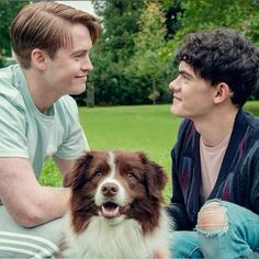 two young men are sitting on the grass with a brown and white dog in front of them