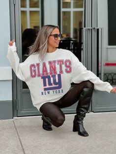 a woman squatting on the ground in front of a building wearing black boots and a giants sweatshirt