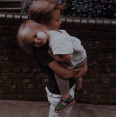 a young boy holding a doll on his back while standing next to a brick wall