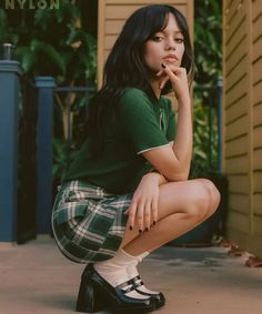 a woman in green shirt and plaid skirt sitting on the ground with her hand under her chin