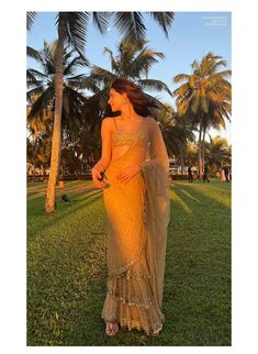 a woman in a yellow sari standing on grass with palm trees behind her and looking off to the side