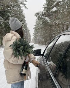 a woman holding a christmas tree next to a parked car in the snow with a bottle of wine