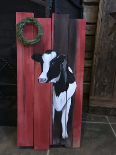 a painting of a black and white cow on a red wooden board with a wreath