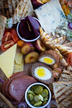 an assortment of meats and cheeses on a cutting board with bread, olives, tomatoes, peppers, eggs, and other foods