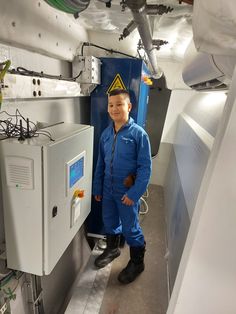 a young boy in blue jumpsuit standing next to an industrial machine and smiling at the camera