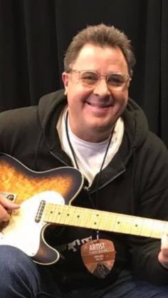 a man with glasses is holding a guitar and smiling at the camera while sitting in front of a black background