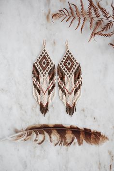 two pairs of beaded earrings with feathers on a marble surface next to fern leaves