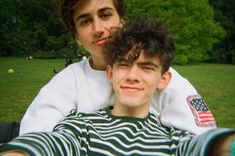 two young men sitting on top of each other in the grass with trees in the background