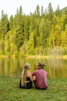 a man and woman sitting on the grass next to a lake with trees in the background