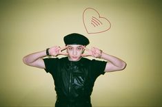 a young man wearing a black hat and holding his hands behind his head with a heart drawn on the wall above him