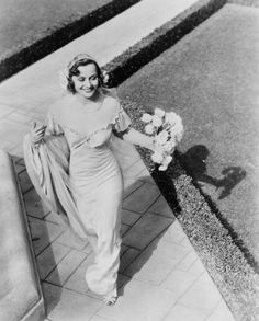 an old photo of a woman walking down the street with flowers in her hair and wearing a dress