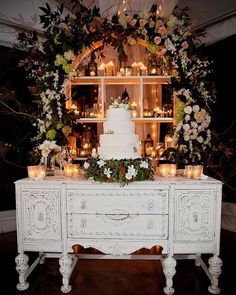 a wedding cake on top of a white table with candles and flowers in the background