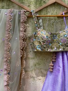 A three-piece tea green and lavender mirror lehenga set from the Priti Sahni collection. This beautiful raw silk lavender lehenga with floral mirror hand embroidery belt detail is paired with a tea green mirror bustier in sequin, beads, and mirror multi-hued hand embroidery. The lehenga has side hanging ball tassels to the waistline. And the blouse has thread-sequins tassel tie-up at the back. This outfit is completed with a tea green tulle mirror work net dupatta with scalloped edging. Bollywood Style Lavender Sharara With Mirror Work, Festive Lavender Sets With Mirror Work, Purple Dola Silk Lehenga With Gota Work, Lavender Mirror Work Dupatta For Party, Party Lehenga With Mirror Work In Lavender, Lavender Lehenga With Mirror Work For Party, Lavender Party Dupatta With Mirror Work, Party Lavender Lehenga With Mirror Work, Traditional Lavender Sets With Mirror Work