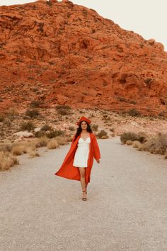 a woman in a white dress and red coat walking down a dirt road with a mountain behind her