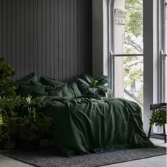 a bed covered in green sheets and pillows next to a window with potted plants