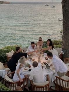 a group of people sitting around a table with food and drinks in front of the ocean