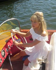 a woman sitting on the steering wheel of a small boat in the water while wearing a white dress