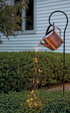 a watering can is lit up with fairy lights in front of a house on the lawn