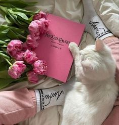 a white cat laying on top of a bed next to flowers and a pink book