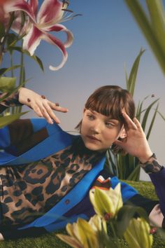 a woman sitting in the grass with her hand on her head and flowers behind her