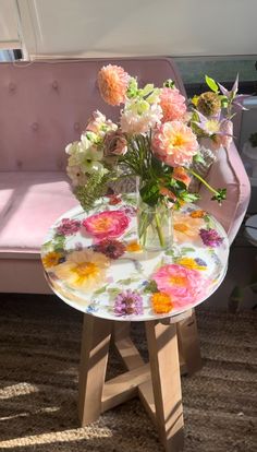 a glass table with flowers on it in front of a pink chair and window sill