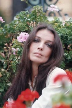 a woman with long brown hair sitting in front of some red and pink flowers, looking at the camera