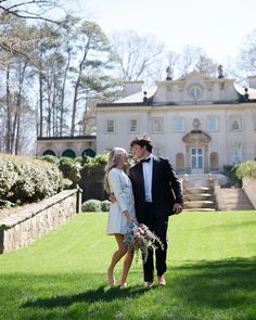 a young man and woman standing in front of a large house with grass on the lawn