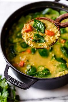 a spoon full of soup with spinach and carrots in it, being held up by a wooden spoon