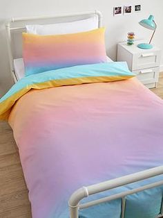 a bed with pink, blue and yellow sheets in a white room next to a lamp