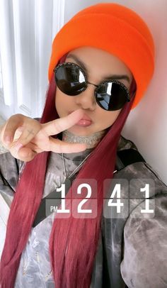 a woman with long red hair wearing sunglasses and an orange beanie pointing at the camera