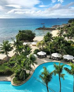 an outdoor swimming pool surrounded by palm trees and the ocean in the background with white umbrellas