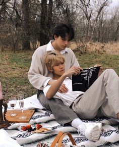 two people sitting on a blanket with food and drinks in front of them, one reading a book