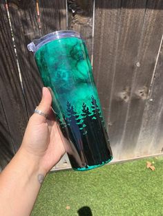 a hand holding a green and black cup with trees painted on the inside, in front of a fence