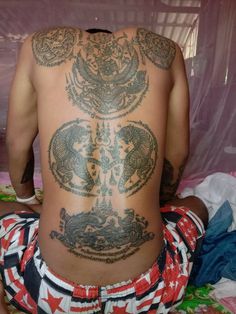 the back of a man with tattoos on his upper and lower body, sitting on a bed