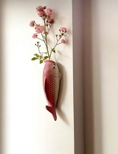 a fish shaped vase with pink flowers hanging on the wall next to a white wall