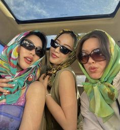 three women sitting in the back of a car wearing scarves and sun visors