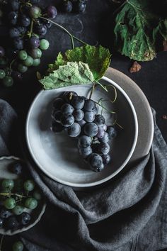 a white plate topped with black grapes next to green leafy leaves on top of a table